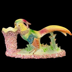 May include: A ceramic figurine of a pheasant with a pink and white vase. The pheasant is painted in shades of green, yellow, red, and blue. The vase is decorated with pink and white flowers.