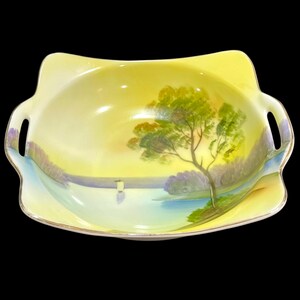May include: A square ceramic bowl with two integrated handles, featuring a hand-painted landscape scene of a tree, a body of water, and a distant shore. The bowl has a yellow background, a gold-trimmed rim, and a smooth, glossy finish.