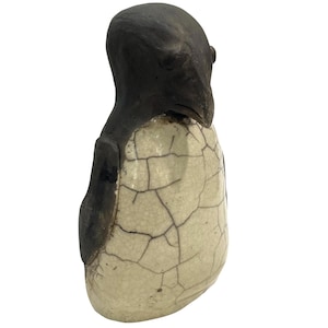 May include: A ceramic penguin figurine featuring a dark charcoal-colored head and flippers contrasted with a cream-colored body. The body displays a distinct crackle glaze finish with dark, intersecting lines across the entire surface area.
