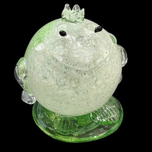 Peut inclure: Figurine de grenouille en verre fantaisiste avec une couronne, corps vert clair et transparent moucheté. La grenouille a des yeux noirs et repose sur une base verte. Objet décoratif.