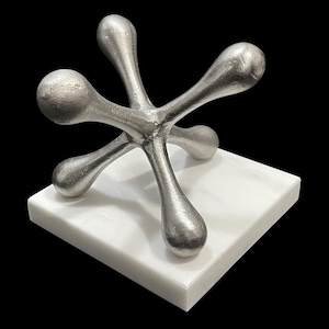 Puede incluir: Una escultura de metal plateado con forma de jack, con cinco brazos redondeados, sobre una base cuadrada de mármol blanco. La escultura tiene una superficie texturizada y está sobre un fondo negro. Esta pieza decorativa añade un toque moderno.