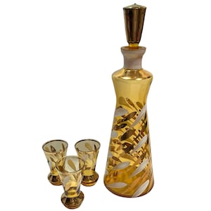 Peut inclure: Un ensemble de carafe doré vintage avec un bouchon assorti et trois verres à liqueur. La carafe et les verres présentent un motif de feuilles blanches et dorées. L'ensemble est probablement en verre.