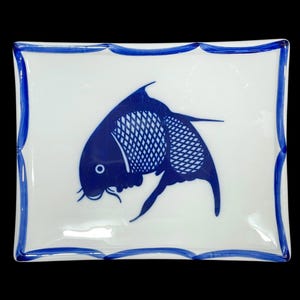 May include: A square, white ceramic plate with a blue fish design. The fish is stylized with a detailed pattern on its body. The plate has a blue scalloped border, creating a decorative edge.