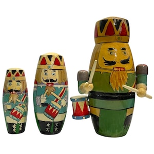 May include: Three cylindrical wooden nutcracker figures of varying sizes, each painted with a yellow face, black moustache, and beard. They wear crowns and green uniforms, holding drumsticks and playing small red and white drums with gold accents.