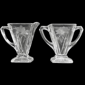 May include: A set of two clear glass sugar and creamer containers with etched floral designs and square bases.
