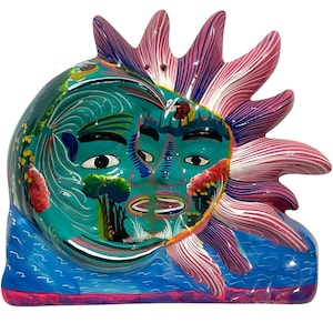 May include: Hand-painted ceramic sun face sculpture. The sun features a teal face with black eyes, a smiling mouth, and colorful details. The sun's rays are pink and white. The base is blue. A unique piece of art.