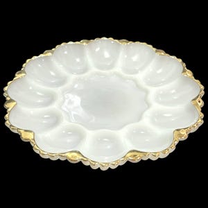 Op de afbeelding: Een ronde eierserveerschaal van wit melkglas met twaalf individuele ovale inkepingen in een cirkel rond een glad middenstuk. De geschulpte buitenrand van de schaal is afgewerkt met een metallic gouden rand.