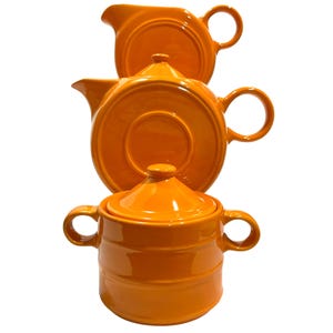 Peut inclure: Un ensemble de trois articles de cuisine en céramique orange vif composé d'un pot à lait, d'une théière avec couvercle et d'un sucrier avec couvercle. Chaque pièce présente une finition lisse et brillante, des anses arrondies et une couleur unie.