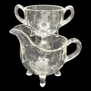 May include: Clear glass creamer and sugar bowl set. The creamer has a spout and three small feet. Both pieces feature etched floral designs. The sugar bowl has two handles. The set is on a black background.