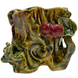 May include: A ceramic planter with a naturalistic design. The planter is glazed in shades of green, brown, and yellow, with a pair of red cherries and a frog figurine. The planter has a textured surface and an organic shape.