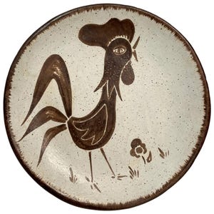 May include: A round ceramic plate with a speckled off-white glaze and a dark brown rim. The centre features a hand-painted dark brown rooster standing next to a small flower. The design uses simple, bold lines to depict the bird's form.