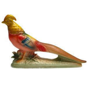 May include: A ceramic figurine of a golden pheasant with a yellow crest, orange and red chest, and blue-tipped wings. The bird stands on a green base with molded leaf details and features a long, tapered tail extending horizontally to the right.