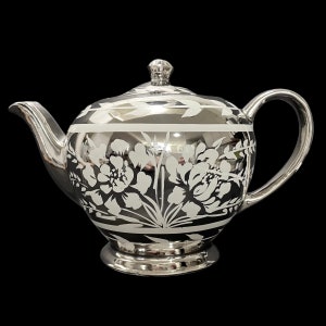 Könnte beinhalten: Eine silberne Teekanne mit einem weißen Blumendesign. Die Teekanne hat einen runden Körper und einen Ausguss an der Seite. Die Teekanne steht auf einem schwarzen Hintergrund.