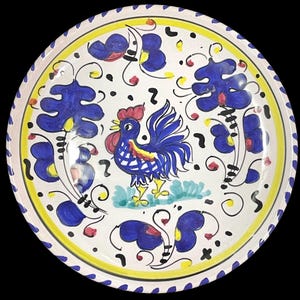 Puede incluir: Un plato redondo de cerámica con un gallo azul pintado a mano en el centro, rodeado de motivos florales azules estilizados y pequeños detalles amarillos. El borde presenta un patrón repetitivo azul y blanco con una fina línea amarilla.