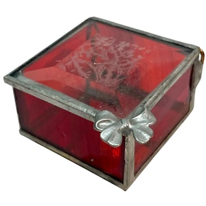 May include: A square box constructed from translucent red glass panels held together by silver-toned metal framing. The lid features an etched floral pattern on the top surface, and a decorative metal bow is attached to one corner of the frame.