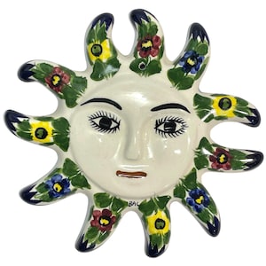 Peut inclure: Une décoration murale en céramique représentant un visage de soleil. Le visage crème a des yeux noirs et une bouche marron. Les rayons du soleil sont décorés de feuilles vertes et de fleurs colorées rouges, jaunes et bleues. Finition brillante.