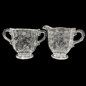 May include: A clear glass creamer and sugar bowl set. Both pieces feature a floral etched design and curved handles. The sugar bowl has two handles, while the creamer has one handle and a spout. The set is on a black background.