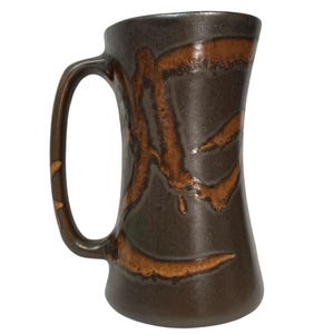 May include: A tall, cylindrical ceramic mug with a dark brown, speckled glaze and a large, curved handle. The exterior features abstract, flowing brushstroke patterns in a lighter, earthy orange-brown tone that wrap around the body.