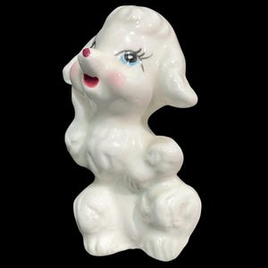 May include: A white ceramic poodle figurine with a glossy finish. The dog has painted blue eyes with long black eyelashes, a pink nose, and a small open mouth showing a pink interior. The poodle is depicted in a seated position with textured fur.
