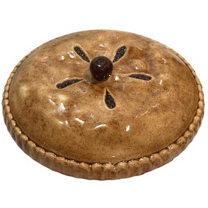 May include: A round ceramic pie dish with a lid, featuring a glossy tan and brown speckled finish that mimics a baked crust. The lid has a central dark brown knob and five decorative steam vents arranged in a star pattern around the center.