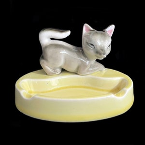 May include: A ceramic ashtray with a gray cat figurine on top. The ashtray is yellow and oval shaped.
