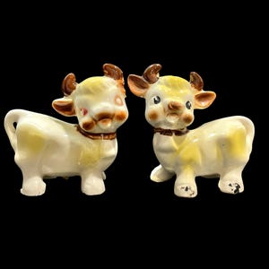 May include: Two ceramic cow figurines. The cows are white with yellow spots and accents, brown horns, and brown collars. The figurines have a vintage, cartoonish style and are set against a black background.