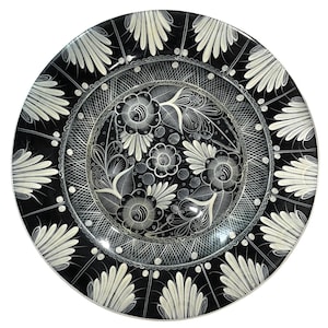 May include: A black decorative plate with a white floral design. The plate features a central floral pattern surrounded by intricate details and a border of stylized white fan-like shapes. The plate is round and has a glossy finish.