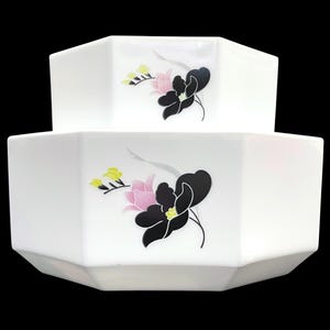 May include: A two-tiered white ceramic bowl with an octagonal shape. Each tier features a floral graphic print consisting of a large black flower, a smaller pink flower, and tiny yellow buds, all arranged on the smooth white surface of the bowl.