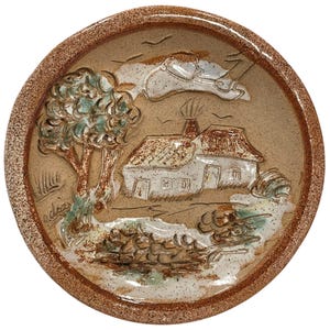 May include: A ceramic plate featuring a raised design of a house, trees, and birds. The plate is brown with white and green accents. The plate is signed "ndrea".
