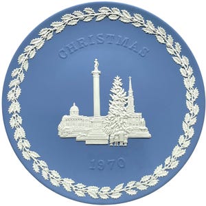 May include: A round light blue ceramic plate featuring a white relief design of a Christmas scene with a tall column, a large tree, and buildings. The word CHRISTMAS is embossed at the top, and the year 1970 is embossed at the bottom center.