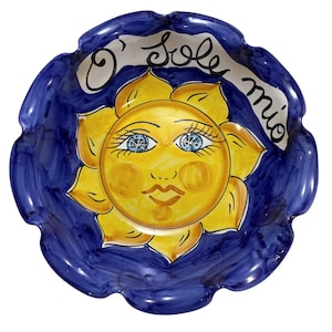 May include: A round, scalloped-edge ceramic bowl featuring a hand-painted yellow sun with a face in the centre. The background is a deep blue with brushstroke textures. The text O' sole mio is written in black script across the upper portion.