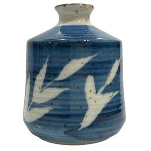 May include: A ceramic vase with a cylindrical body and a narrow neck, featuring a deep blue glaze with horizontal ridges and white leaf-shaped patterns. The surface is decorated with small brown speckles throughout, and the rim is white.