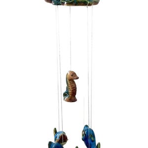 May include: A ceramic wind chime featuring a seashell, a seahorse, and four dolphins. The seashell is green, brown, and blue. The seahorse is brown. The dolphins are blue. The wind chime is hanging from a string.