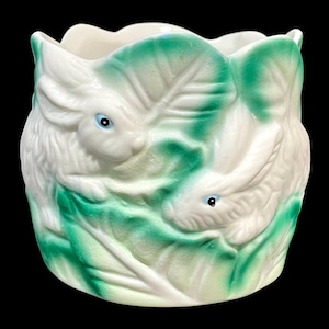 Könnte beinhalten: Ein weißer Keramik-Pflanztopf mit gewelltem Rand, der geprägte weiße Hasen zeigt, die sich zwischen grünen Blättern befinden. Die Hasen haben blaue Augen. Der Pflanztopf ist ein dekorativer Artikel.