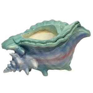 Op de afbeelding: Een keramische schelpvormige container in tinten blauw, teal en roze. De container heeft een textuur en een gedetailleerd spiraalontwerp aan het puntige uiteinde. De opening is afgezet met een lichtere kleur.