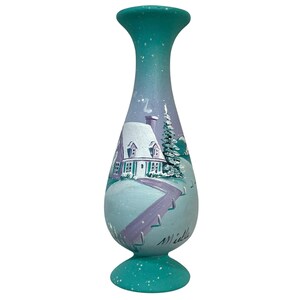 May include: A teal vase with a flared rim and base, featuring a hand-painted winter scene of a house with a snow-covered roof, a tall evergreen tree, and a purple path. The surface is decorated with white speckles and the signature Mielle.