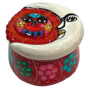May include: A hand-painted ceramic trinket box with a lid featuring a red sun and a white crescent moon. The box is red with floral designs in pink, green, and blue. The sun has a smiling face with colourful details.