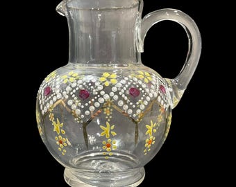 May include: A clear glass jug featuring a hand-painted design of white dotted diamond patterns, yellow floral accents, and small red circular motifs. The jug has a rounded body, a wide mouth with a pouring spout, and a curved glass handle.