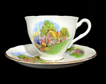 May include: A white porcelain teacup and saucer set featuring a colorful illustration of a thatched-roof cottage surrounded by lush green trees and vibrant flowers. The rim of both the cup and the saucer is finished with a thin gold metallic trim.