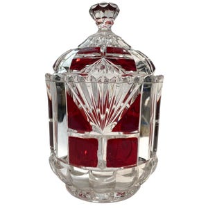 Puede incluir: Un tarro de cristal tallado transparente con tapa, con un interior rojo vibrante. El tarro tiene un diseño geométrico con un patrón de estrella en la tapa y acentos rojos rectangulares. La base es festoneada.