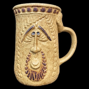 以下が含まれることがあります： 顔のデザインが施されたベージュのセラミックマグ。マグにはハンドルが付いており、目、鼻、ひげを含む彫刻された顔で装飾されています。マグはテクスチャ加工された表面で、おそらく手作りです。