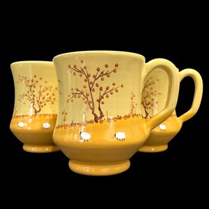 May include: Three yellow ceramic mugs with a rural design. Each mug features a brown tree with small brown dots, white sheep, and a yellow landscape. The mugs have a curved handle and a wide base.