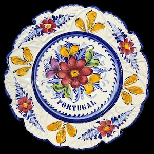 May include: A white ceramic plate with a blue rim and a floral design in shades of red, yellow, green, and purple. The plate has a scalloped edge and the word "PORTUGAL" written in blue on the bottom.