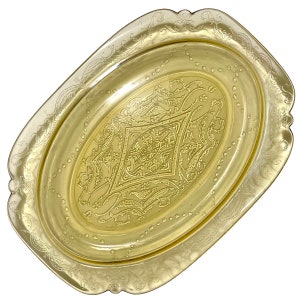 Puede incluir: Una bandeja ovalada de color amarillo con detalles en relieve. La bandeja presenta un diseño central en forma de diamante y un borde decorativo. Los bordes tienen un diseño festoneado. La bandeja es probablemente de vidrio o un material similar.