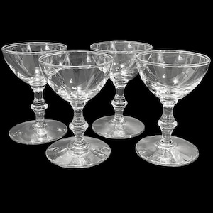 May include: Four clear, stemmed champagne coupe glasses. Each glass has a wide, shallow bowl, a decorative stem, and a round base. The glasses are arranged on a black background.