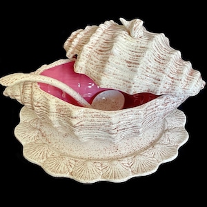 May include: A white ceramic seashell-shaped serving bowl with a lid and a matching serving spoon. The bowl has a pink interior and a speckled brown glaze. The bowl is sitting on a white ceramic plate with a scalloped edge.