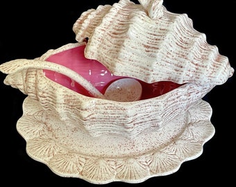 May include: A ceramic tureen shaped like a large conch shell with a matching lid, a ladle, and an underplate. The exterior features a cream-colored glaze with brown speckles, while the interior of the tureen and the ladle bowl are a vibrant pink.