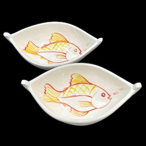 May include: Two white, leaf-shaped ceramic dishes. Each dish features a hand-painted fish design in yellow and red. The fish have a grid pattern on their bodies. The dishes have small handles on each side and are set against a black background.