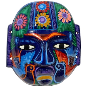 May include: A hand-painted ceramic mask in dark blue. The mask depicts a stylized face with large eyes, a prominent nose, and a closed mouth. It is decorated with colourful floral and bird motifs in shades of orange, blue, green, and purple.