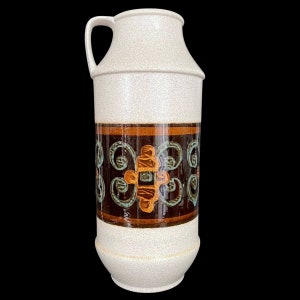 May include: A tall ceramic pitcher with a speckled off-white finish and a single handle. The center features a wide, dark brown horizontal band decorated with repeating orange and teal scrollwork patterns that wrap around the cylindrical body.